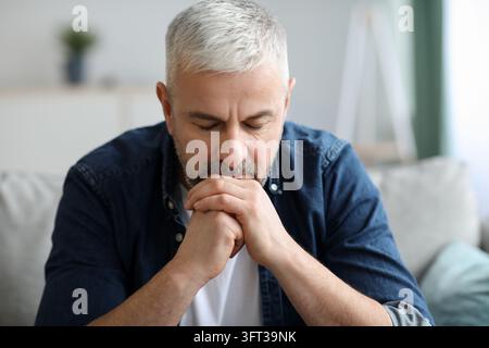 Pensive senior bearded man sitting on sofa in living room at home, leaning on his hands and looking down, having financial problems or thinking about Stock Photo