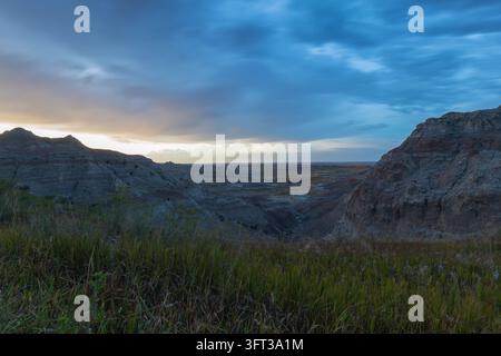 Soft twilight glowing on the horizon as blue storm clouds sweep over eroded buttes and a deep canyon framed by prairie grass, Badlands National Park, Stock Photo