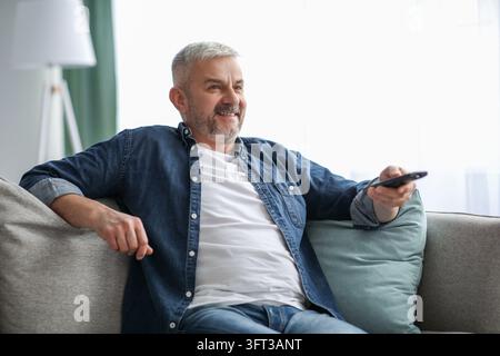 Happy bearded senior man in casual clothes relaxing on couch at home, holding remote, watching TV, copy space. Relaxed matured man having break on sof Stock Photo