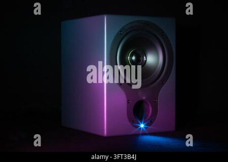 A modern silver gray studio monitor speaker angled on a dark surface illuminated by a dramatic neon purple and cyan ambient glow with a brilliant blue Stock Photo