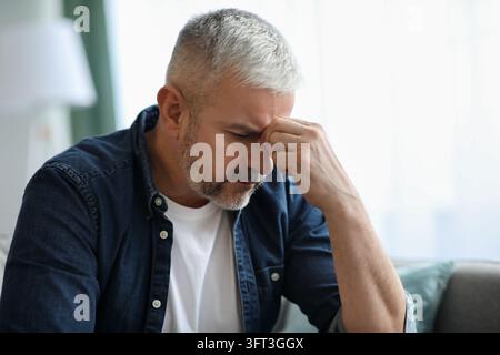 Exhausted matured grey-haired bearded man touching his nose bridge, sitting on couch with closed eyes, home interior. Senior man feeling sick, sufferi Stock Photo