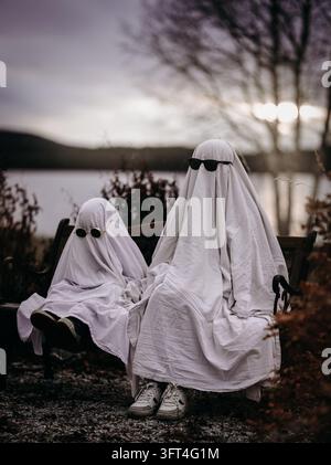 Two people dressed as white sheet ghosts wearing sunglasses sit together on a bench beside a lake at sunset. Playful Halloween concept. Stock Photo