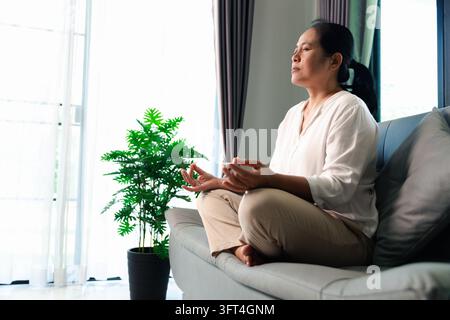 A woman sits cross-legged on a gray sofa with both hands forming meditation-style gestures, beside a leafy potted plant and bright window, leaving ope Stock Photo