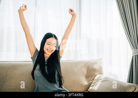 Cheerful woman sitting stretches with raised arms on sofa after relaxing. Lifestyle of a happy freelancer in a luxury living room. Wellbeing joy and c Stock Photo