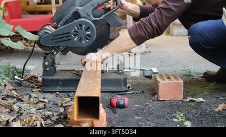 A worker secures a rusty metal profile pipe onto the bed of a cut-off saw, holding the tool by its handle before beginning to cut. Stock Photo