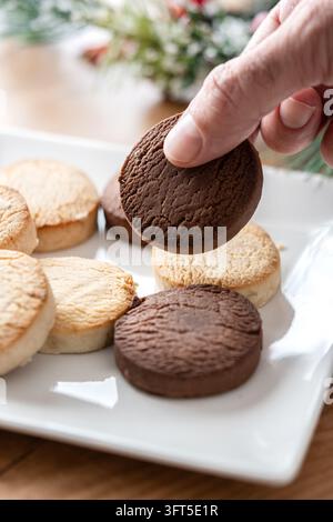 Close-up of a hand picking up a chocolate shortbread cookie from a white plate filled with assorted vanilla and chocolate cookies. Homemade sweet bisc Stock Photo