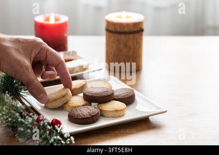 Close-up of a hand picking up a chocolate shortbread cookie from a white plate filled with assorted vanilla and chocolate cookies. Homemade sweet bisc Stock Photo