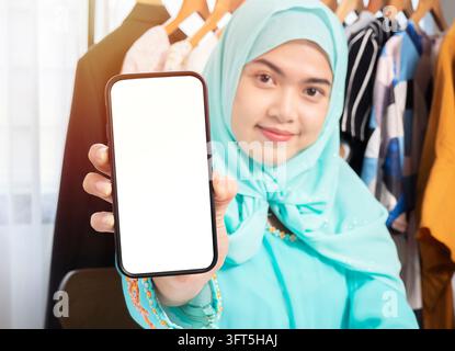 Excited beautiful Asian woman in green sweater and hijab showing blank screen mobile phone isolated over her shop selling through ecommerce Stock Photo