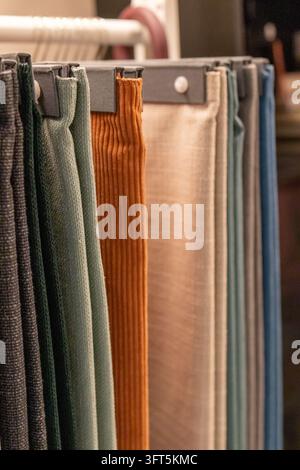 Orange corduroy, beige linen, green and blue upholstery fabric swatches hang vertically from gray holders on a rail in a furniture store. Concept of c Stock Photo