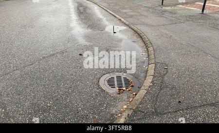 Round metal storm drain grate beside curved curb on wet asphalt road with puddles and fallen autumn leaves Stock Photo