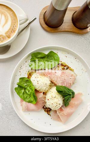 healthy breakfast consists of zucchini fritters, poached eggs, slices of ham and spinach leaves on a white plate and a cup of cappuccino. A laconic Stock Photo