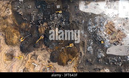 Aerial view of multiple yellow excavators and cranes working on a large muddy construction site with piles of earth and concrete foundations in India. Stock Photo