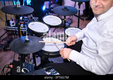 A male drummer in a white shirt holds drumsticks over an electronic drum kit during live entertainment at a ball. Stock Photo