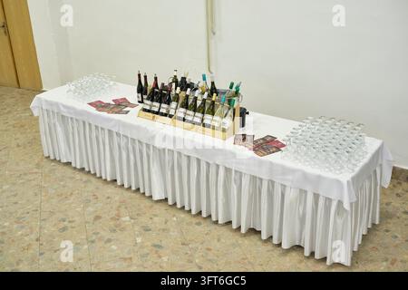 Wine bottles in wooden crates and rows of empty stemmed glasses are arranged on a long white-skirted table for a ball. Stock Photo