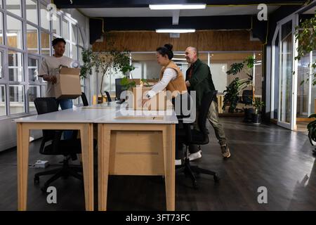 Diverse coworkers moving cardboard boxes and arranging plants on communal desks in modern office Stock Photo