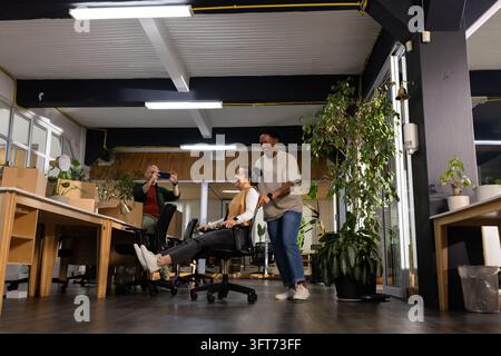 Diverse coworkers pushing woman sitting in rolling chair in open-plan office, taking phone picture Stock Photo