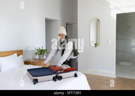 Adult female packing carry-on with rust clothing on bed in modern bedroom, wearing beanie and scarf Stock Photo