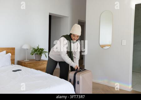 Asian woman in beanie bending, handling hardshell suitcase in bedroom, copy space Stock Photo