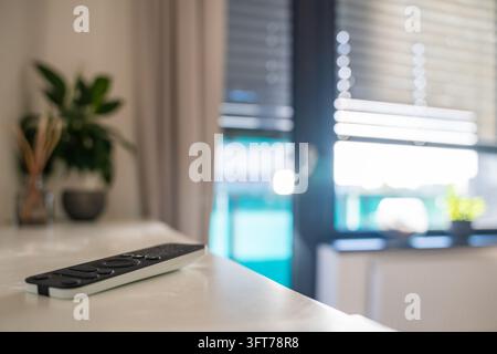 Remote control for motorized window blinds lies on a shelf with blurred blinds in the background. Smart home automation allows convenient daylight Stock Photo