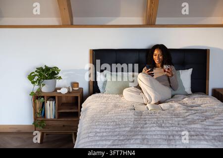 African American woman sitting on bed at home in light gray loungewear holding smartphone and glass Stock Photo
