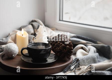 Black cup of hot drink on wooden tray with burning candle, pine cone, white Christmas tree candle and cozy blanket beside snowy window. Warm winter mo Stock Photo