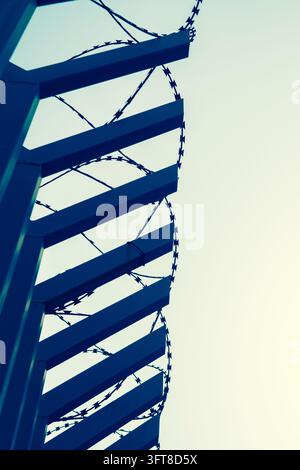 Blue metal slats and coils of barbed wire form a stark security barrier against a pale sky in Beirut, Lebanon. Stock Photo