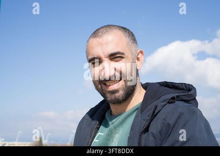 A smiling bearded man in a dark jacket poses outdoors beneath a partly cloudy sky in Lebanon. Stock Photo
