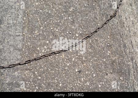 A rusty metal chain stretches diagonally across rough grey pavement, creating a barrier against the textured ground. Stock Photo