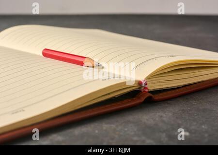 red pencil on open lined notebook on dark desk, blank yellow paper and brown leather cover book in soft focus with copy space, creative writing Stock Photo