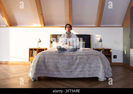 Woman sitting cross-legged on king-size bed in loft bedroom, wearing hoodie and checking smartwatch Stock Photo