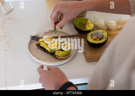 Woman cook sprinkling chopped herbs over open-faced avocado toast near halved avocado on counter Stock Photo