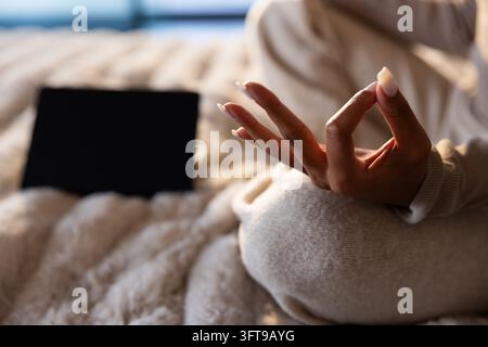 African American woman sitting cross-legged on plush bed in loungewear holding mudra, nails, tablet Stock Photo