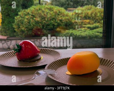 An eye-level indoor photograph capturing two modern artisanal mousse pastries placed on disposable paper plates on a wooden table. On the left, a real Stock Photo