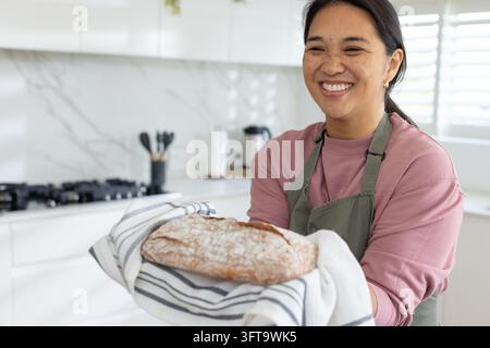 Asian woman smiling holding baked loaf wrapped in striped towel, wearing green apron in kitchen Stock Photo