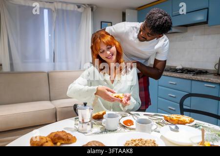 Young man gives his smiling redhead girlfriend a shoulder massage as they share breakfast in a cozy modern kitchen, relaxed and affectionate weekend m Stock Photo