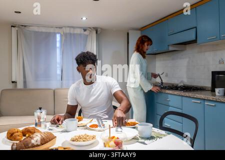 Diverse young couple starting their morning routine by sharing a healthy breakfast, with the man eating at the table and the woman preparing food in t Stock Photo