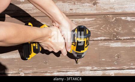 Hands operate a yellow and black cordless power drill against weathered wooden boards, with a second battery powered tool resting nearby in an overhea Stock Photo
