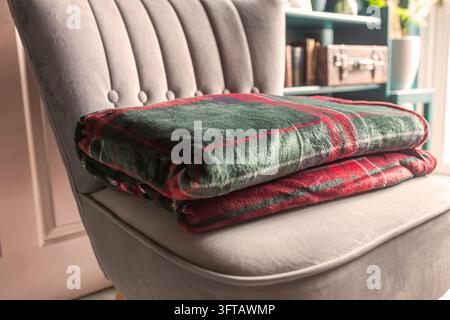 Green red tartan fleece blanket throw folded on a grey chair in a dark green and gold room, with sunlight shining into the room. Stock Photo