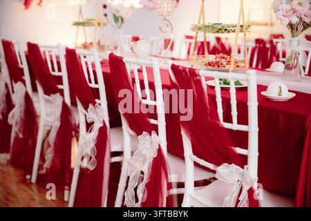 Elegant Wedding Reception Table Setup with Red Draped Chairs and White Tiffany Chairs Stock Photo