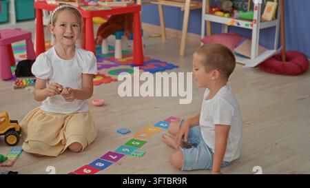 Girl and brother sit and kneel on a wooden floor arranging numbered cards and toy truck beside a shelf and puzzle mat in building; playful bonding. Stock Photo