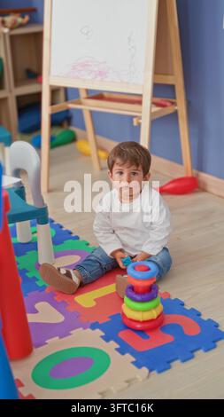 Boy stacking colorful plastic rings on a foam puzzle mat, holding a blue ring and reaching with hand near an easel in building; curiosity learning. Stock Photo