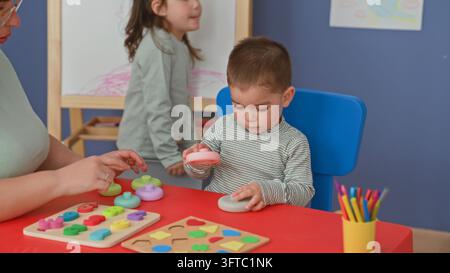 Woman helps toddler stack colorful rings and grip toy on red table in preschool building; concentration learning. Stock Photo