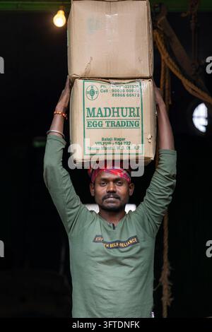 A worker in a green shirt and red head cloth balances boxes on his head inside a dark warehouse lit by a bulb, West Bengal, Howrah, India Stock Photo