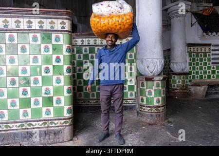 A smiling man in a blue sweater carries a large bag of orange marigolds on his head at the flower market, West Bengal, Kolkata, India Stock Photo