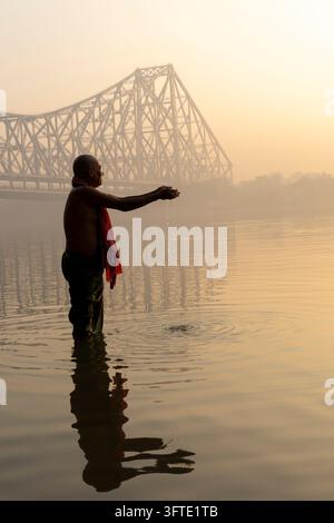 Bather in a red scarf stands waist deep and offers water from his cupped hand in front of a hazy steel bridge, West Bengal, Kolkata, India Stock Photo