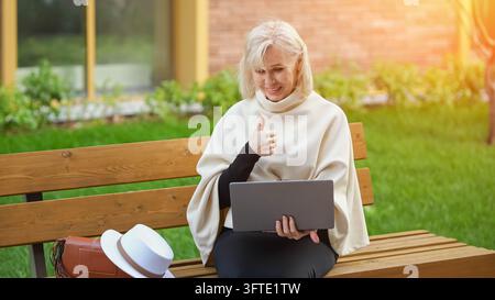 Smiling older woman gives thumbs up while holding laptop on garden bench. Happy computer user presents positive feedback outdoors, with sunlit Stock Photo