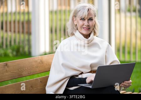 Happy older woman types on laptop while sitting outdoors on wooden bench. Laptop user combines comfortable clothing with remote work, creating relaxed Stock Photo