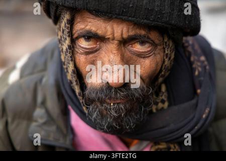 An older man with a gray beard and weathered skin looks intensely at the camera in a black knit hat and dark scarf, West Bengal, Kolkata, India Stock Photo