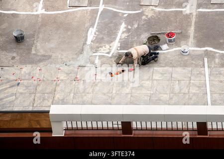 Top view of professional tiler laying ceramic floor tiles on rooftop using adhesive mortar, spacers and leveling tools during renovation. Stock Photo