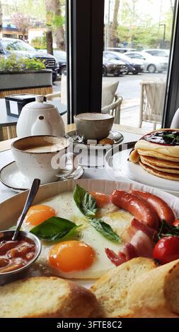 A hearty morning breakfast on a wooden table featuring fried eggs, sausages, tomatoes, bread, pancakes, and a hot cup of coffee at a cafe. Stock Photo